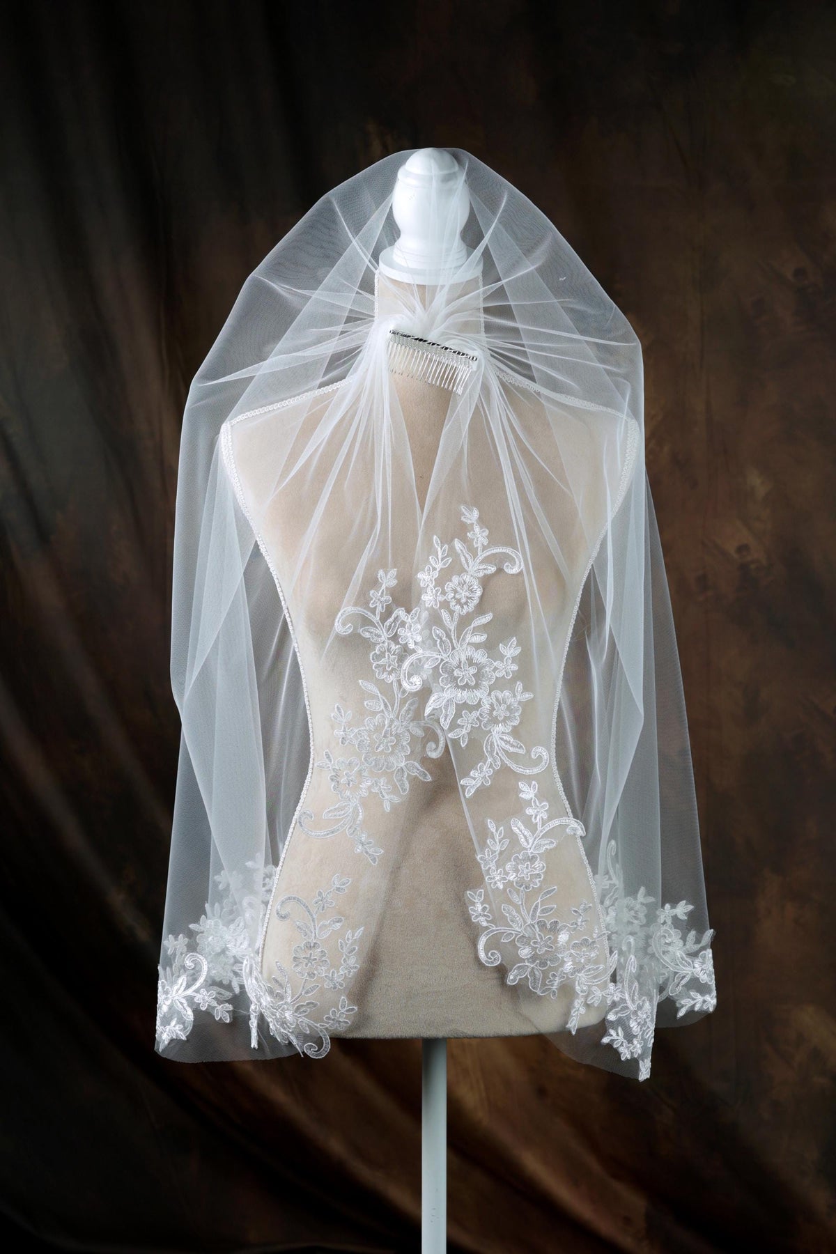 A white wedding veil with intricate lace detailing is displayed on a mannequin against a dark background.