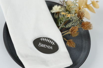 A white napkin with a black oval-shaped name tag that says " Brenda" is placed on a black plate, with some dried flowers and yellow flowers nearby.