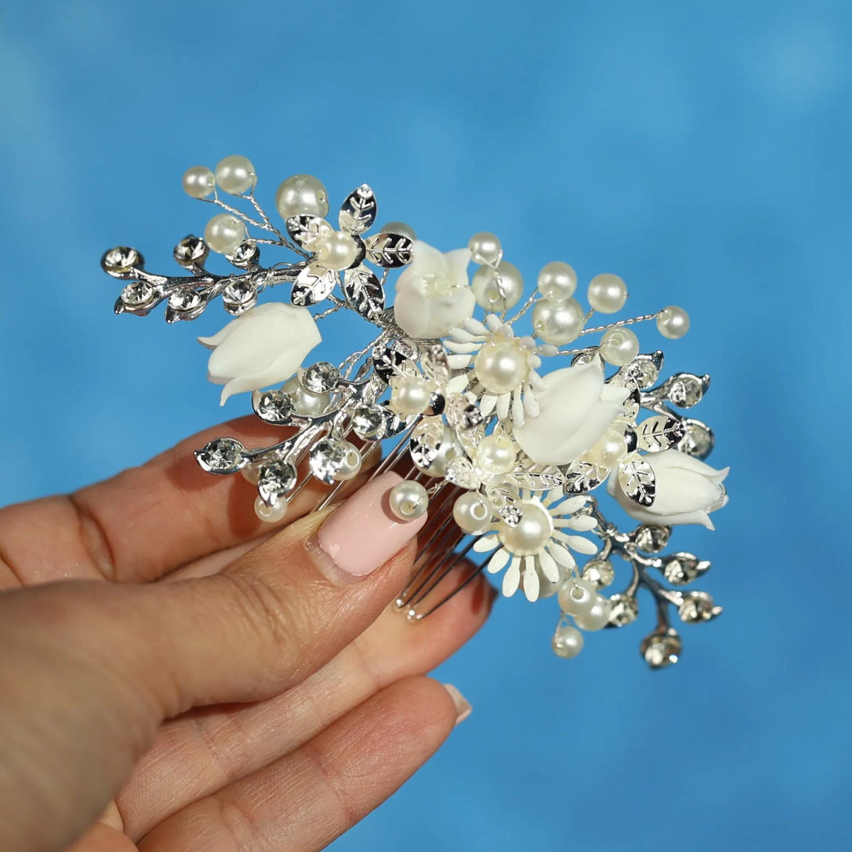 A hand holding a decorative hair accessory with white flowers and pearls against a blue background.