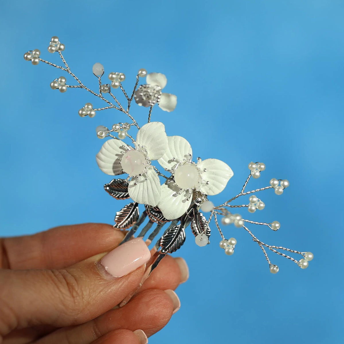 A hand holding a decorative hair clip with white flowers and silver beads against a blue background.