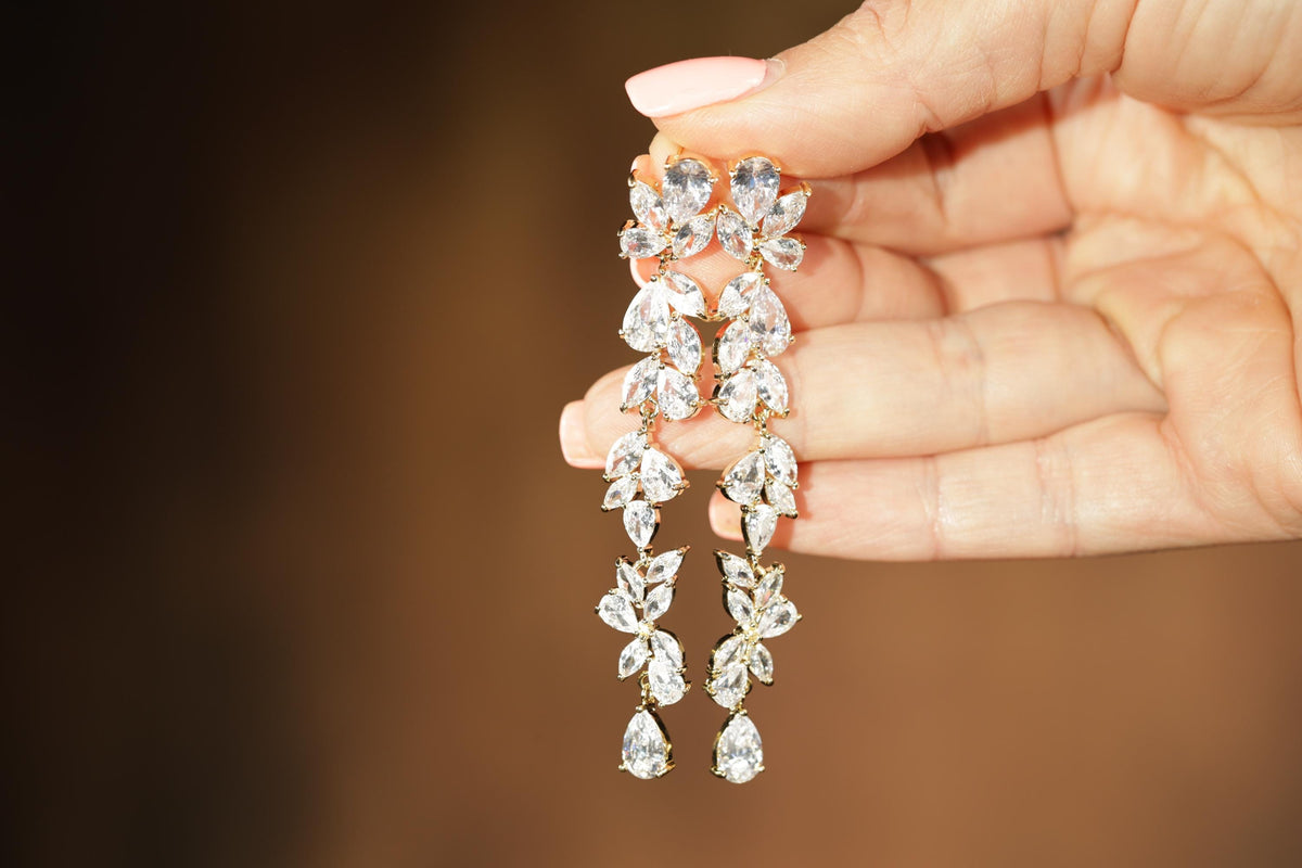 A hand holding a pair of elegant diamond earrings with multiple tiers of clear gemstones.