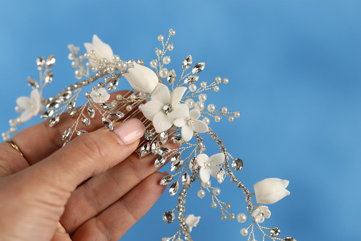 A hand holding a decorative hair accessory with white flowers and silver beads against a blue background.