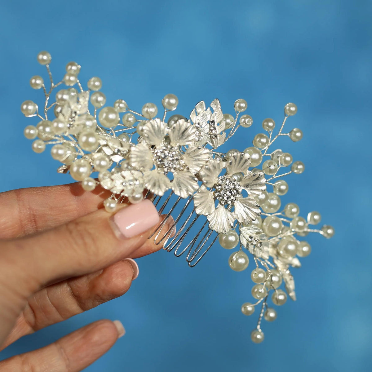 A hand holding a decorative hair accessory with white flowers and pearls against a blue background.