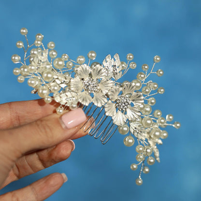 A hand holding a decorative hair accessory with white flowers and pearls against a blue background.