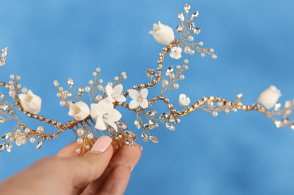 A hand holding a decorative hair accessory with white flowers and gold beads against a blue background.