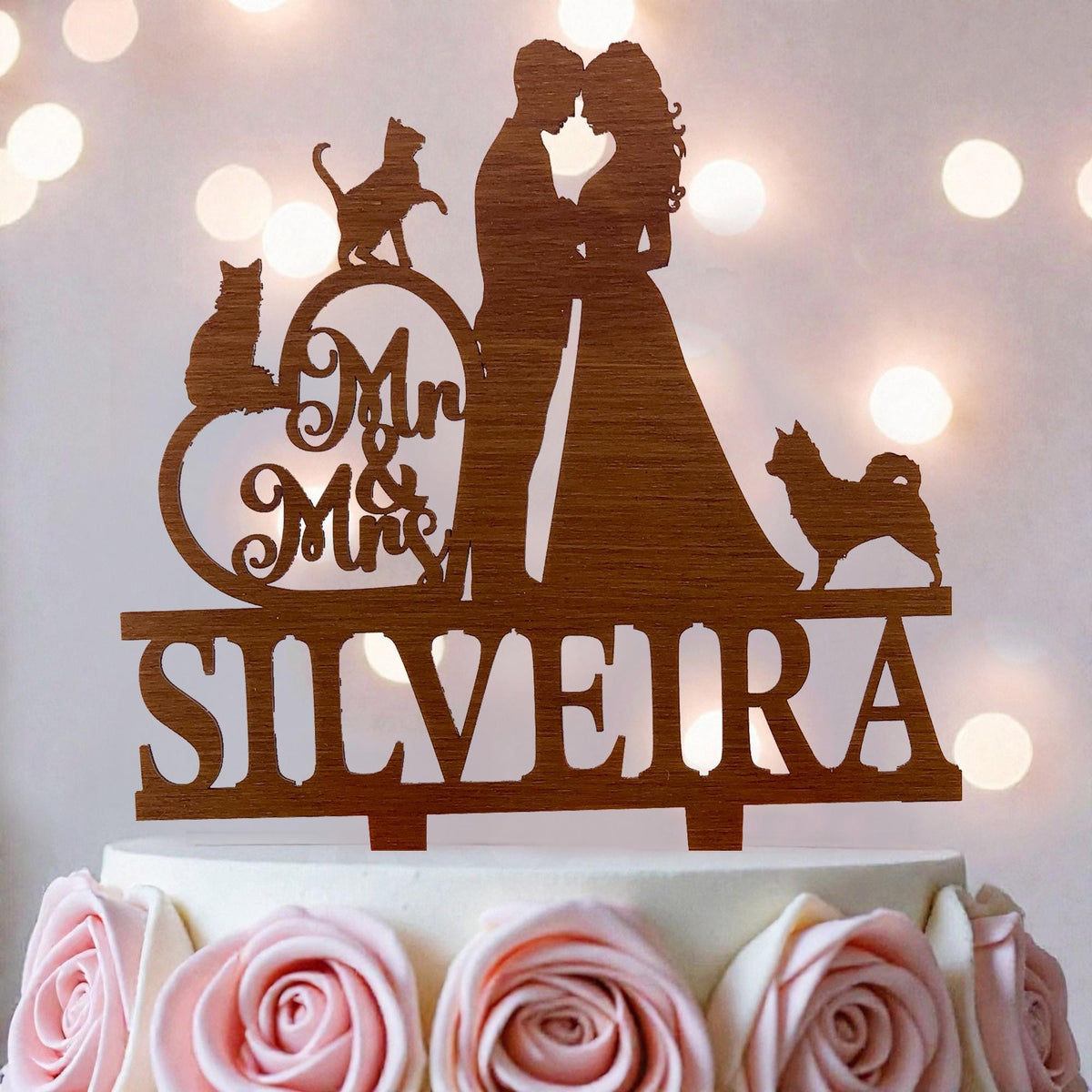 A wooden wedding cake topper with silhouettes of a bride and groom, along with a cat and a dog, and the name "SILVEIRA" written on it.