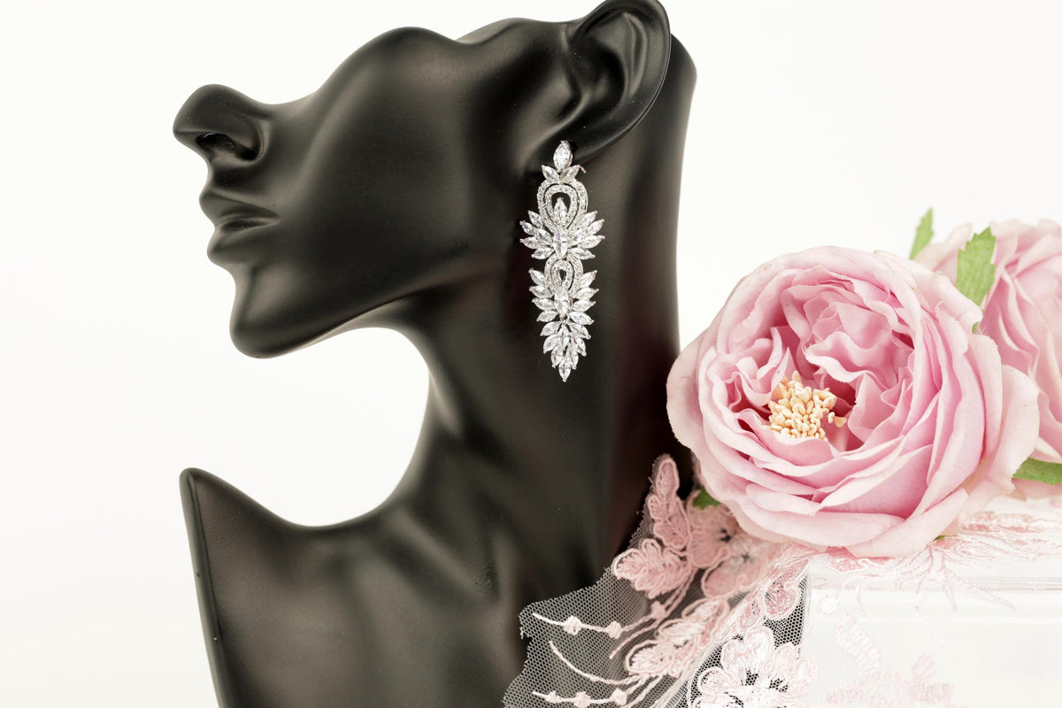 A black mannequin head wearing a pair of elegant diamond earrings, with a delicate pink rose placed next to it.