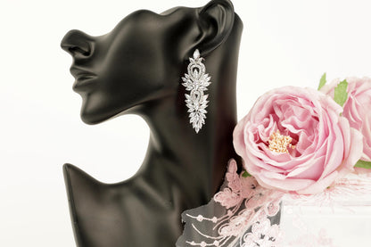 A black mannequin head wearing a pair of elegant diamond earrings, with a delicate pink rose placed next to it.
