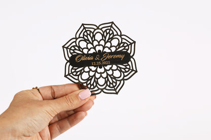 A hand holding a decorative paper cutout with the names 'Olivia & Jeremy' and the date '12.25.2025' printed on it.