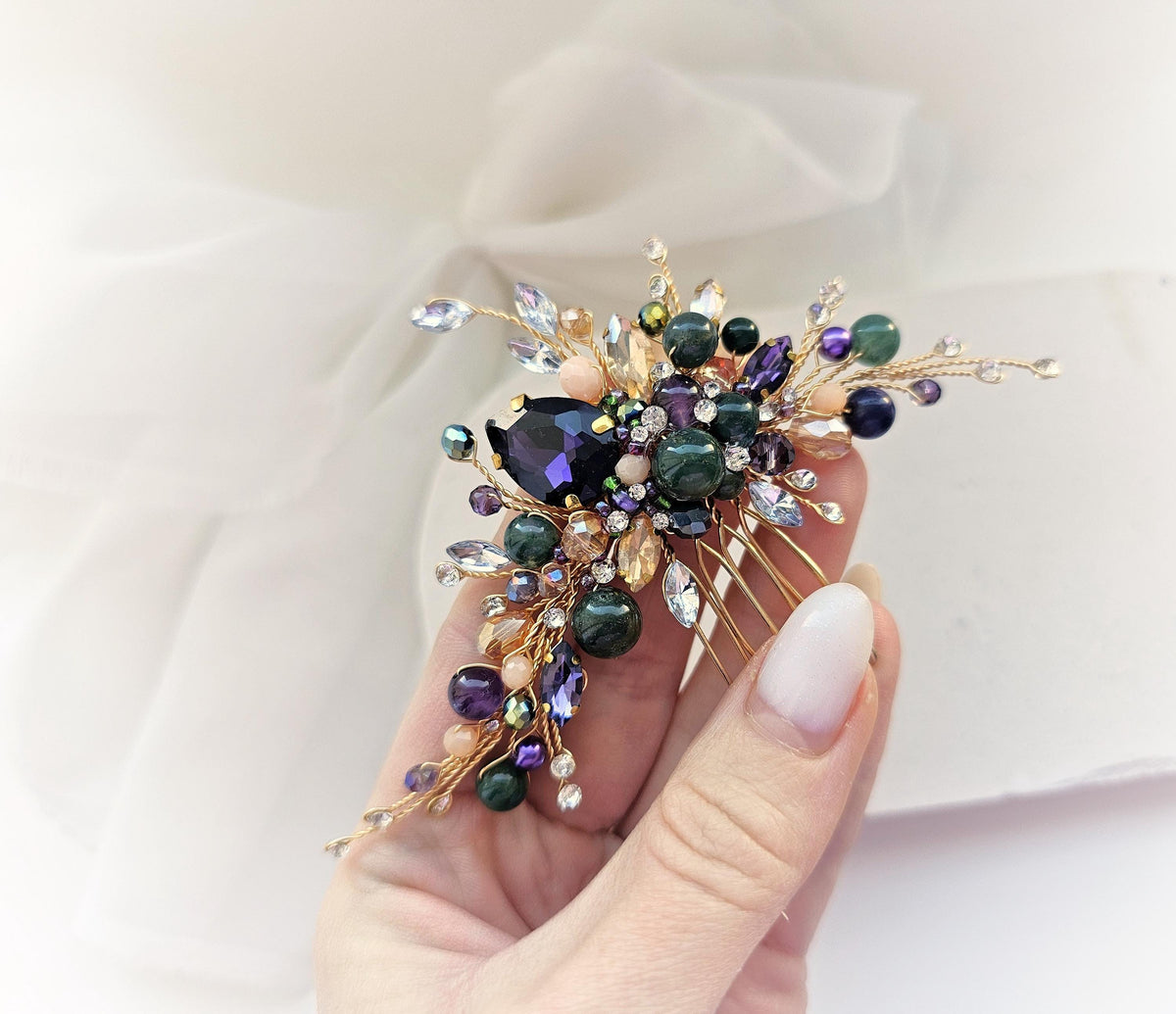 A hand holding a decorative piece of jewelry, which appears to be a hair accessory or brooch. The jewelry is adorned with various gemstones and metallic elements, creating a visually striking and elegant design.