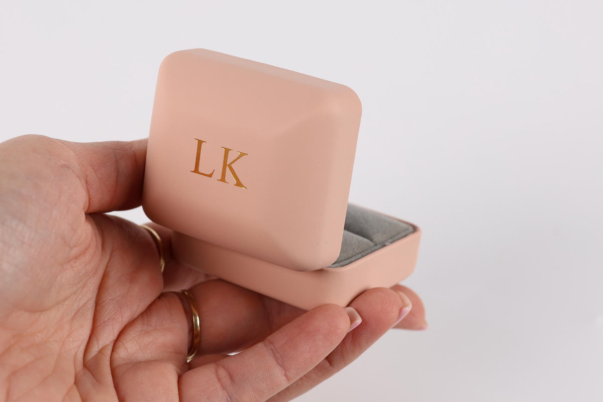 A hand holding a pink jewelry box with the initials "LK" on it.