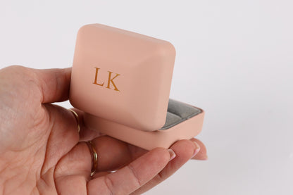 A hand holding a pink jewelry box with the initials "LK" on it.