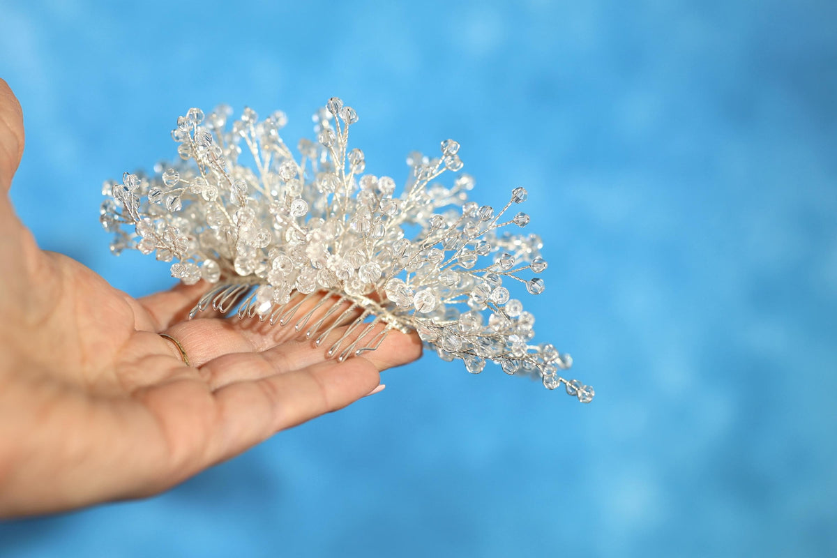 A hand holding a decorative hair accessory made of small, clear beads or crystals against a blue background.