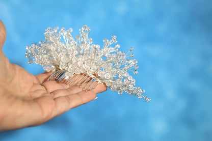 A hand holding a decorative hair accessory made of small, clear beads or crystals against a blue background.