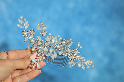 A hand holding a decorative hair accessory with a floral and leaf-like design, featuring small white and clear gemstones or pearls.