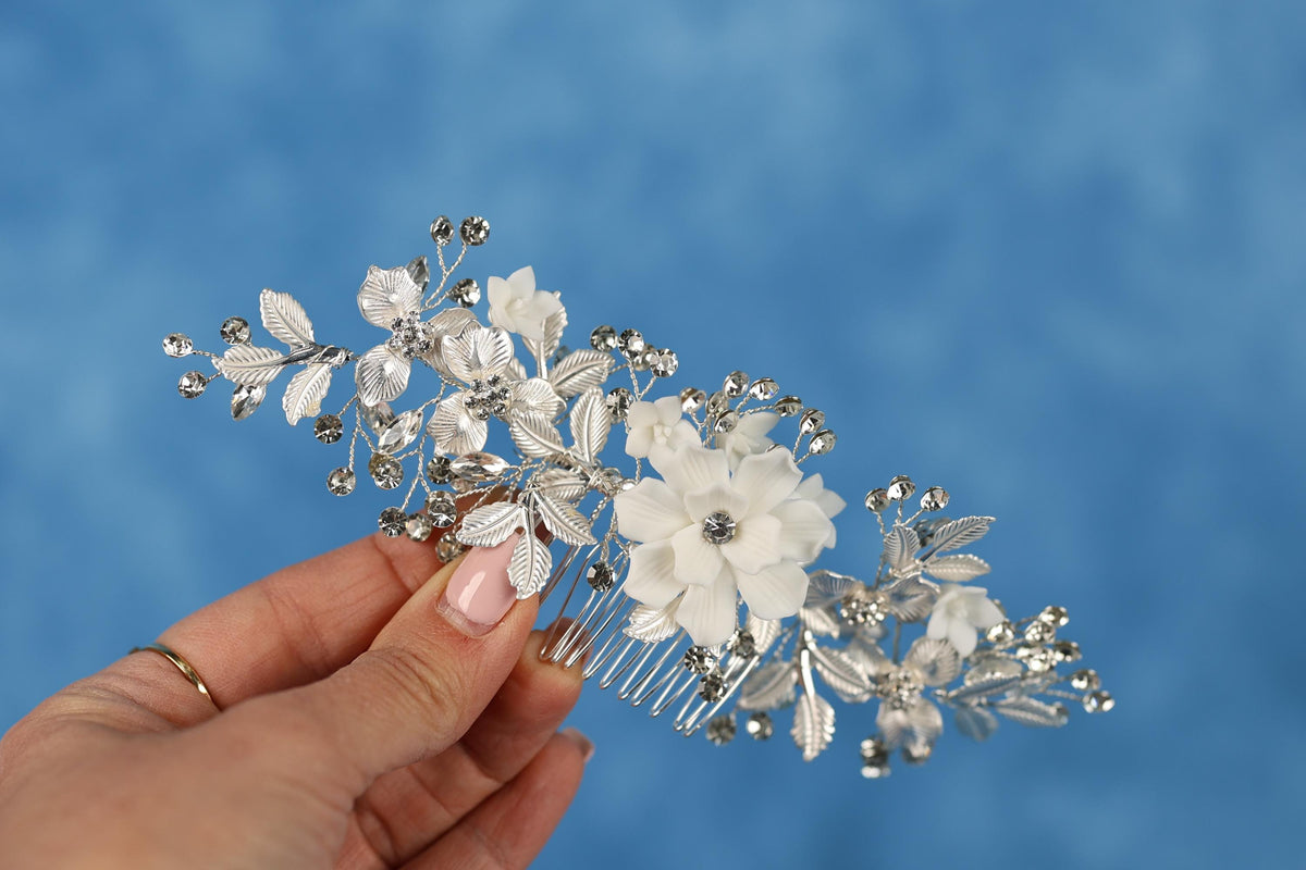 A hand holding a decorative hair accessory with white flowers and silver embellishments against a blue background.