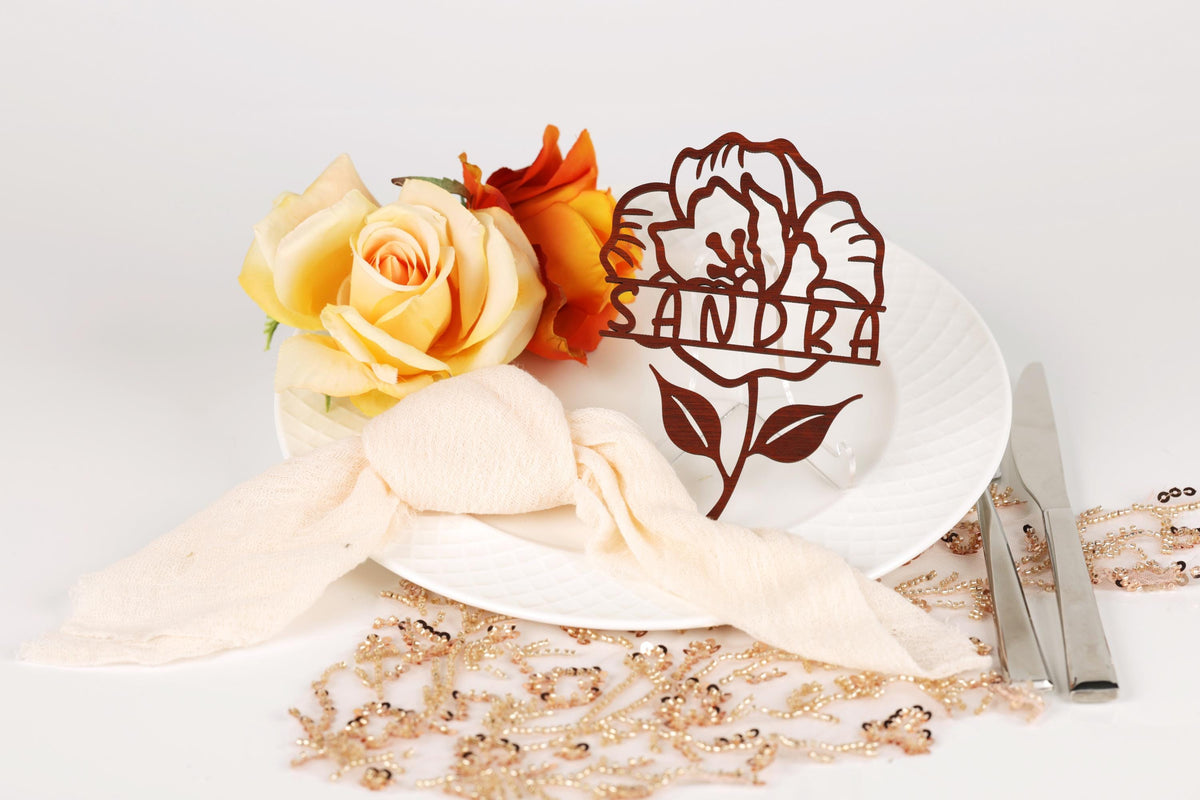 a white plate with a floral design, a ribbon, and a nameplate with a flower design. There are also some flowers and a beaded necklace on the table.