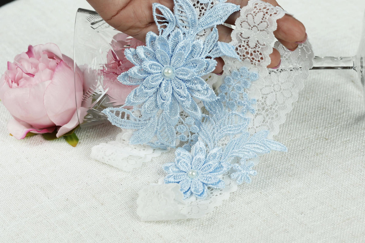 A hand holding a piece of blue lace with white flowers, placed next to a pink rose on a white surface.