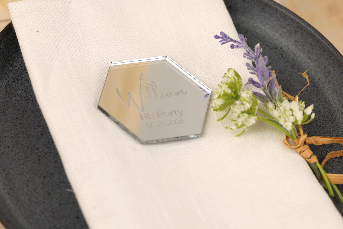 A silver hexagonal plate with a personalized nameplate on it, placed on a white napkin with a small bouquet of purple and white flowers.