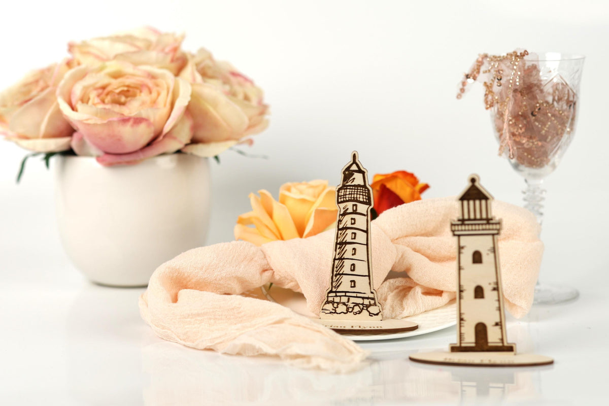 a white vase with pink roses, a wooden lighthouse, a glass of water, and a pink napkin with a wooden lighthouse on it, all arranged on a white surface.
