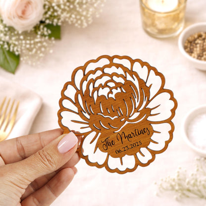 A hand holding a wooden card with a floral design and text on it, with a table setting and other decorative items in the background.