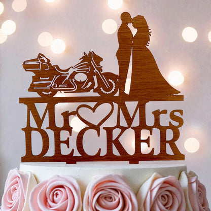 A wooden wedding cake topper with the names 'Mr. and Mrs. Decker' carved into it, along with a silhouette of a couple embracing a motorcycle.