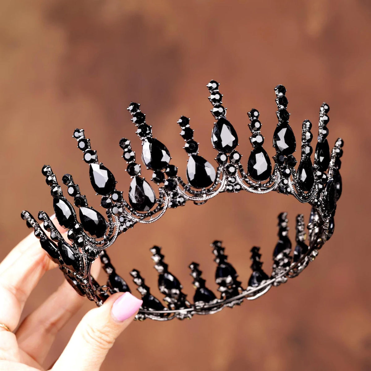 A hand holding a black ornate crown adorned with black gemstones against a brown background.