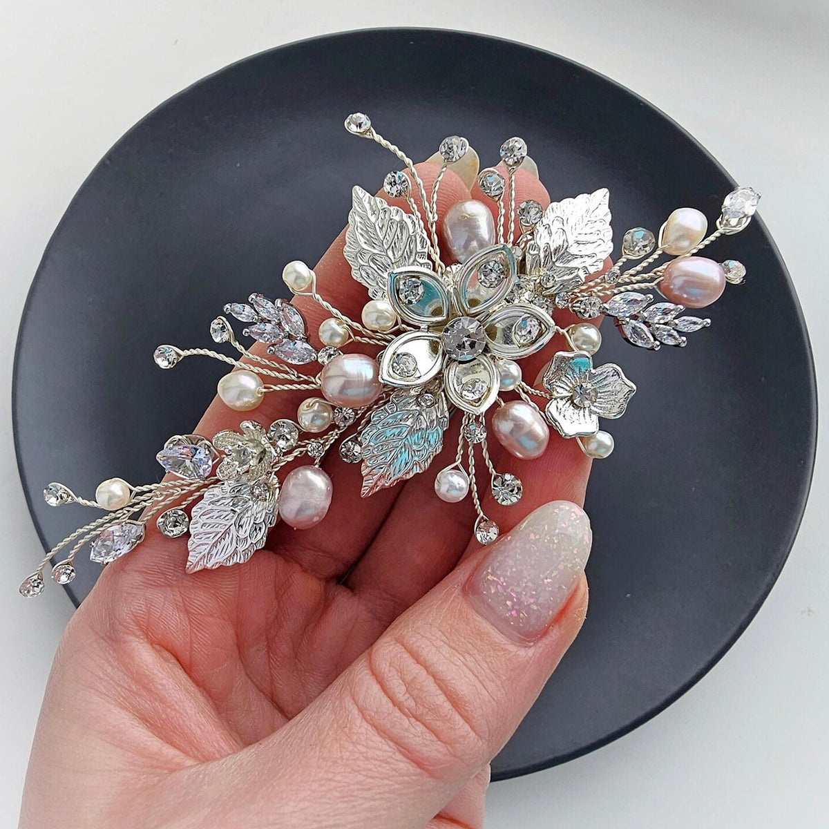 A hand holding a decorative hair accessory with various embellishments, including pearls, crystals, and leaves, against a black background.