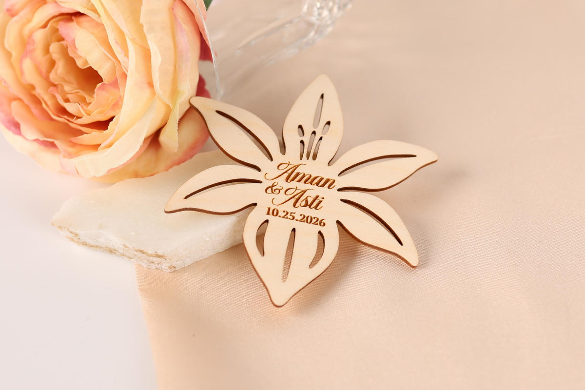 A wooden flower-shaped wedding favor with the names "Aman & Aaliyat" engraved on it, placed next to a pink rose on a light-colored fabric.