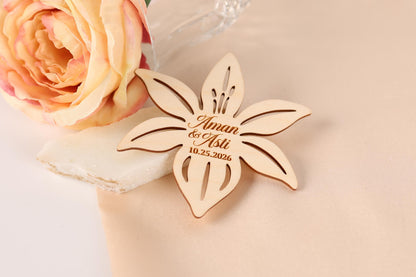 A wooden flower-shaped wedding favor with the names "Aman & Aaliyat" engraved on it, placed next to a pink rose on a light-colored fabric.