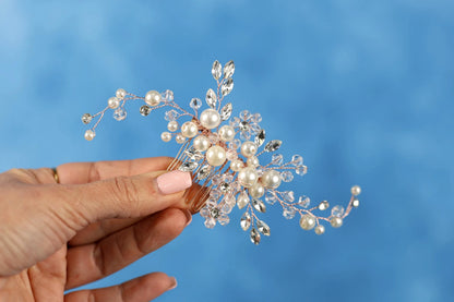 A hand holding a decorative hair accessory made of small pearls and crystal beads against a blue background.