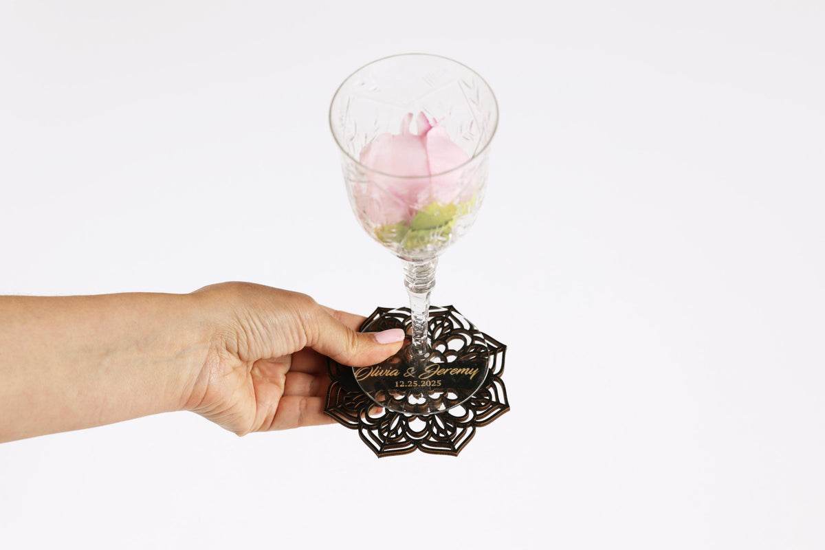 A hand holding a glass with a pink flower in it, and a coaster with the names 'Olivia & Gabriel' printed on it.