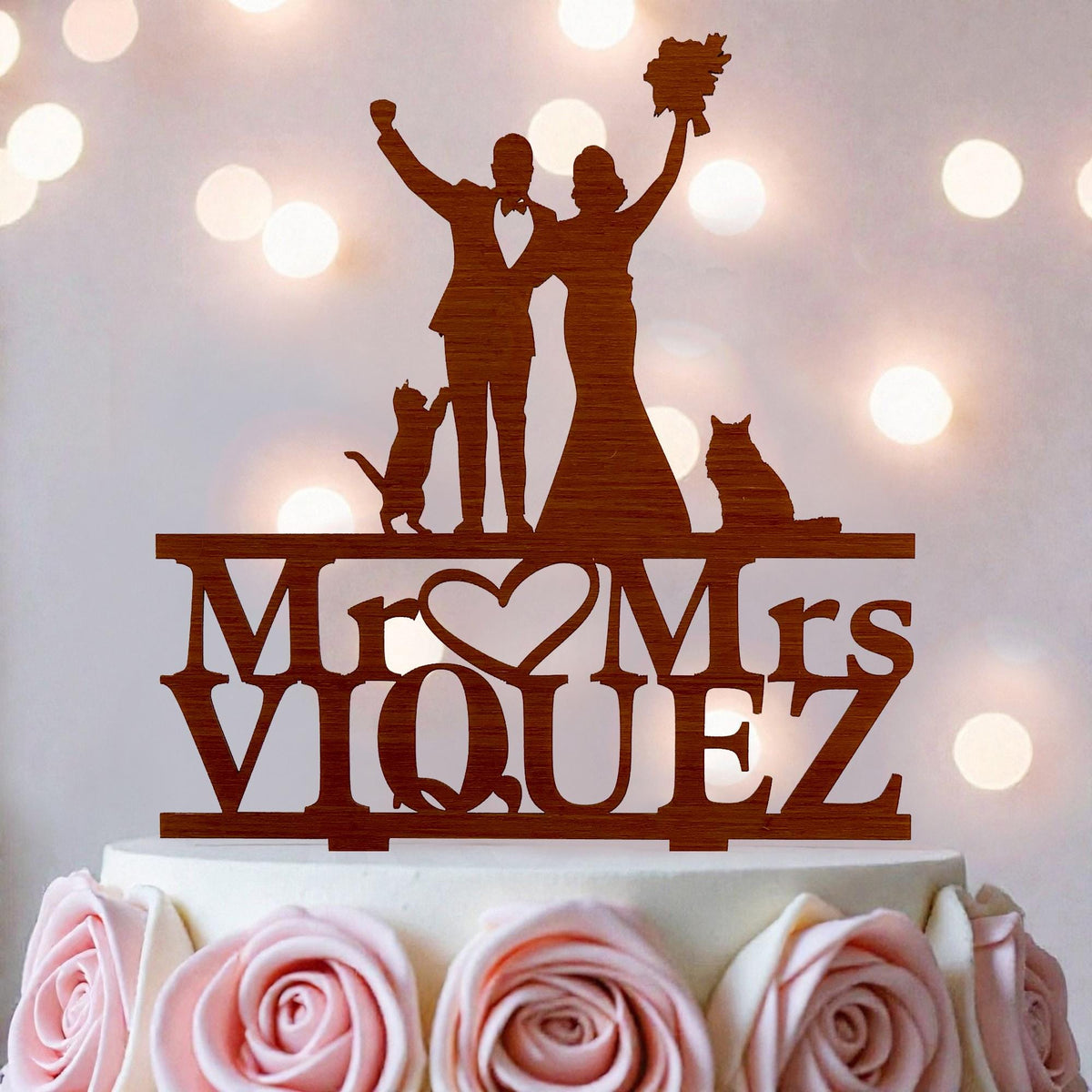 A wooden wedding cake topper with silhouettes of a bride and groom, along with a cat and a rose, and the name "Mr. Mrs. Viquez" written on it.