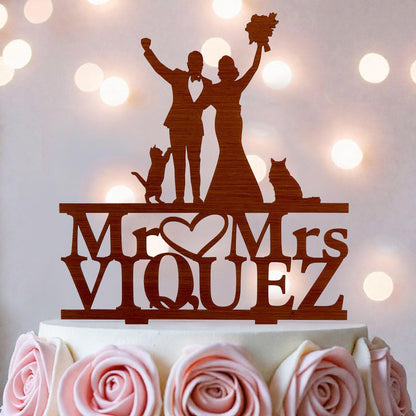 A wooden wedding cake topper with silhouettes of a bride and groom, along with a cat and a rose, and the name "Mr. Mrs. Viquez" written on it.