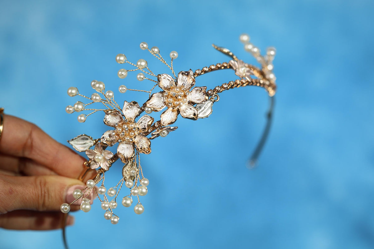 A hand holding a decorative gold-colored tiara adorned with small pearls and floral elements against a blue background.