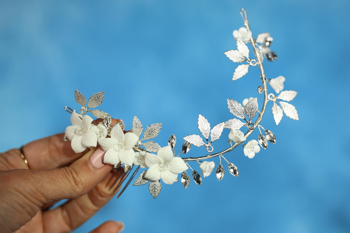 A hand holding a decorative silver hair clip with white flowers and leaves against a blue background.