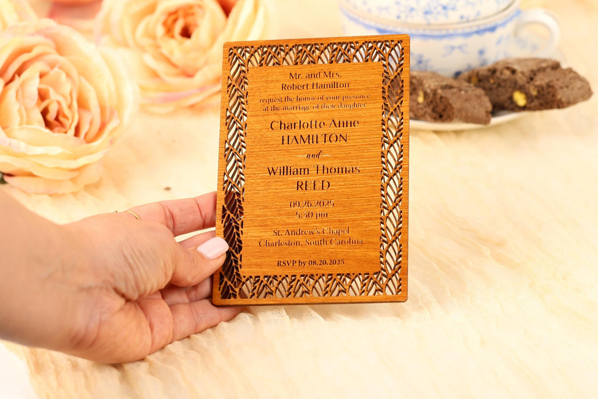 A hand holding a wooden wedding invitation card with text and names on it, placed on a table with flowers and a teacup in the background.