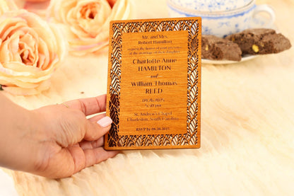 A hand holding a wooden wedding invitation card with text and names on it, placed on a table with flowers and a teacup in the background.