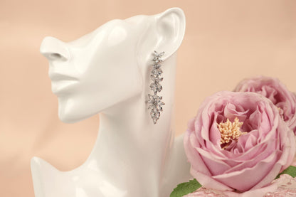 A white mannequin head wearing a pair of dangling earrings with a floral design, placed next to a pink rose.