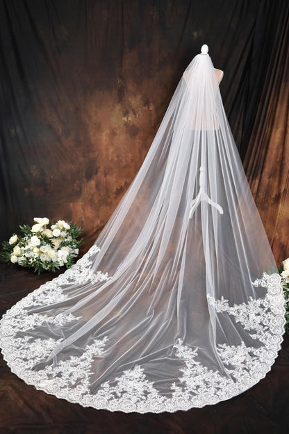 A white wedding veil with a long train and lace trim, displayed against a dark background with a floral arrangement.