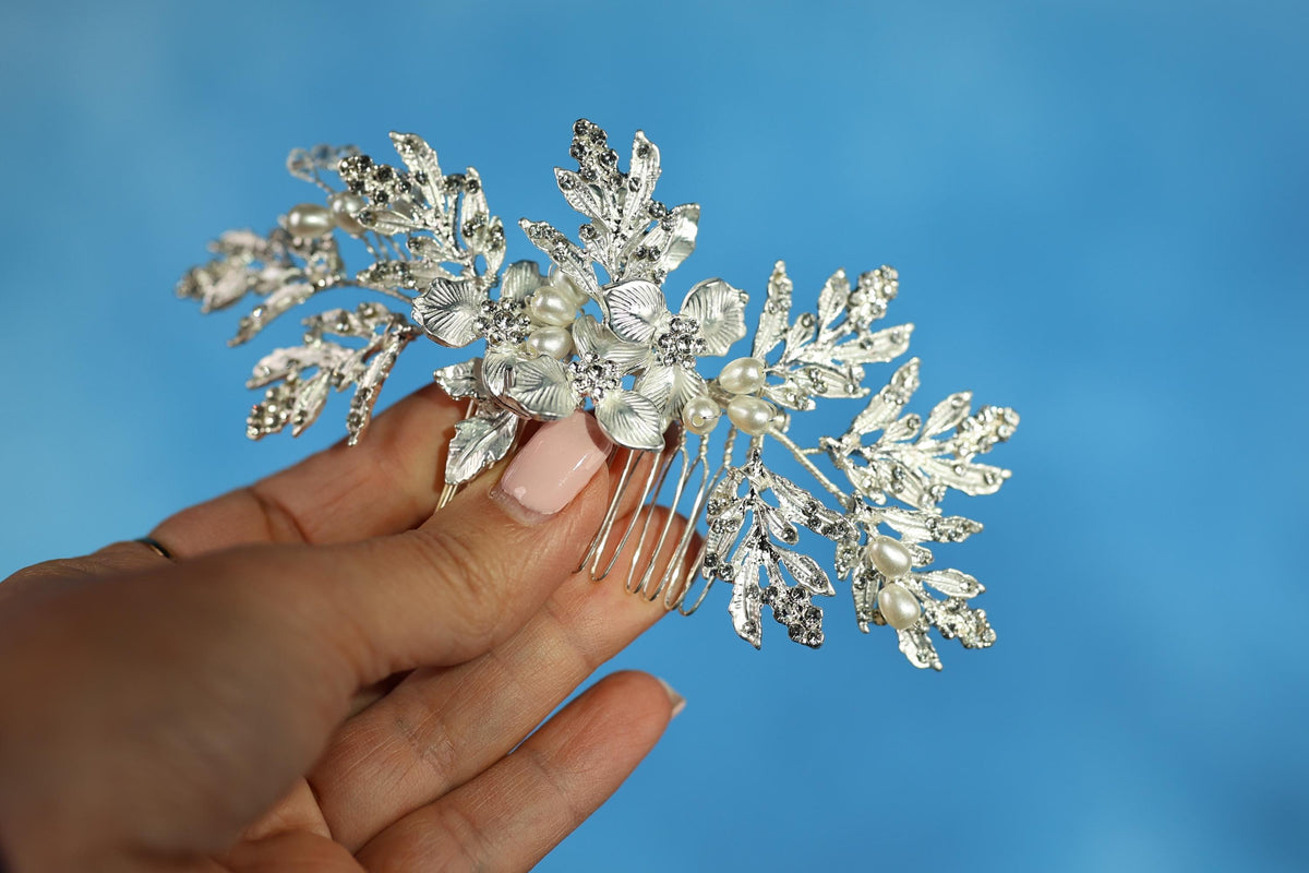 A hand holding a decorative hair accessory with silver and white floral elements against a blue background.