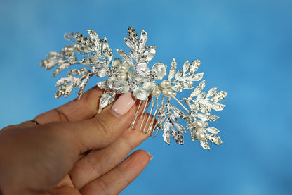 A hand holding a decorative hair accessory with silver and white floral elements against a blue background.