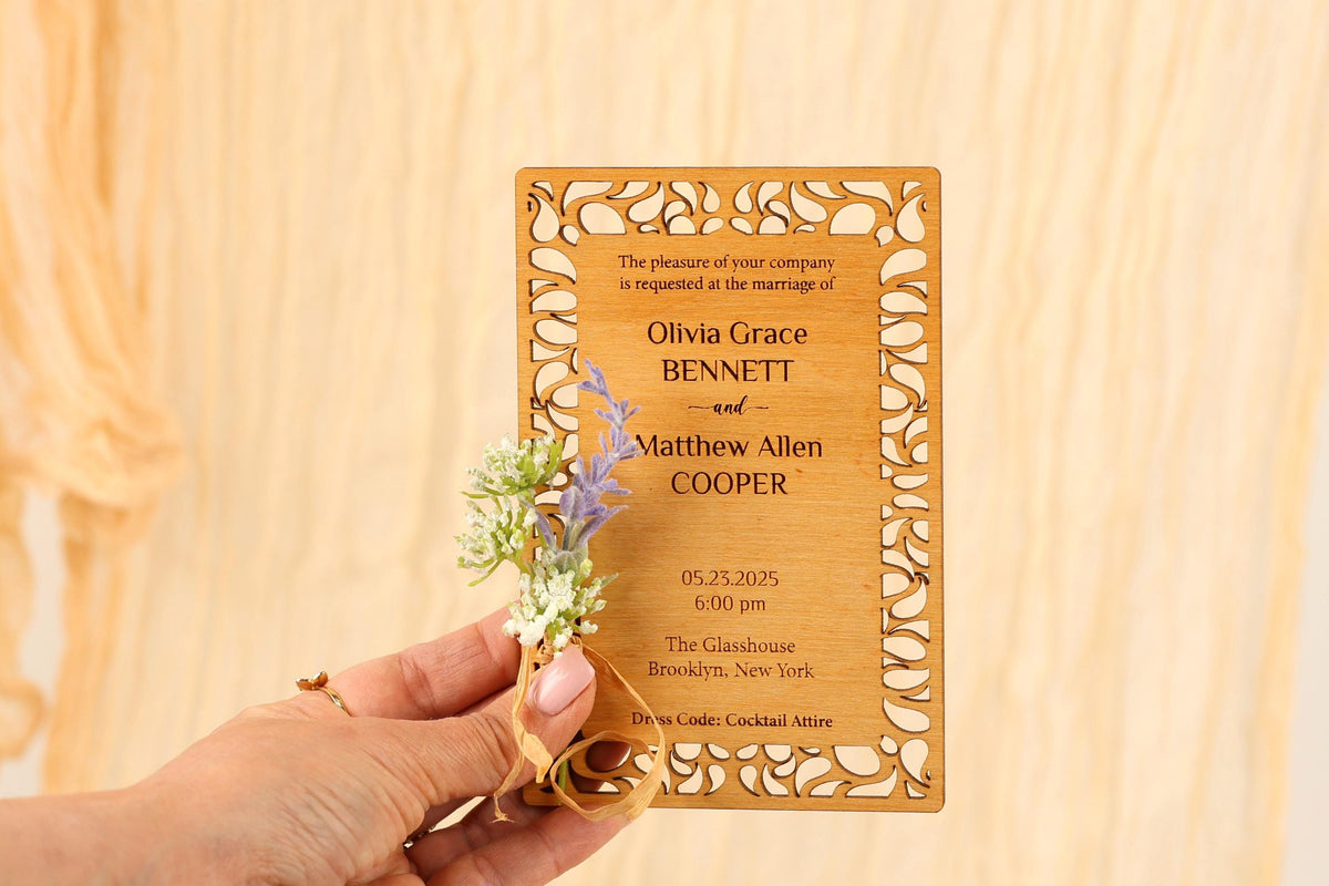 A person's hand holding a wooden wedding invitation card with floral accents.
