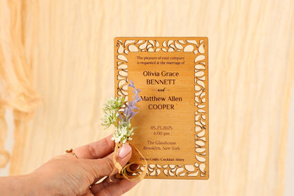A person's hand holding a wooden wedding invitation card with floral accents.