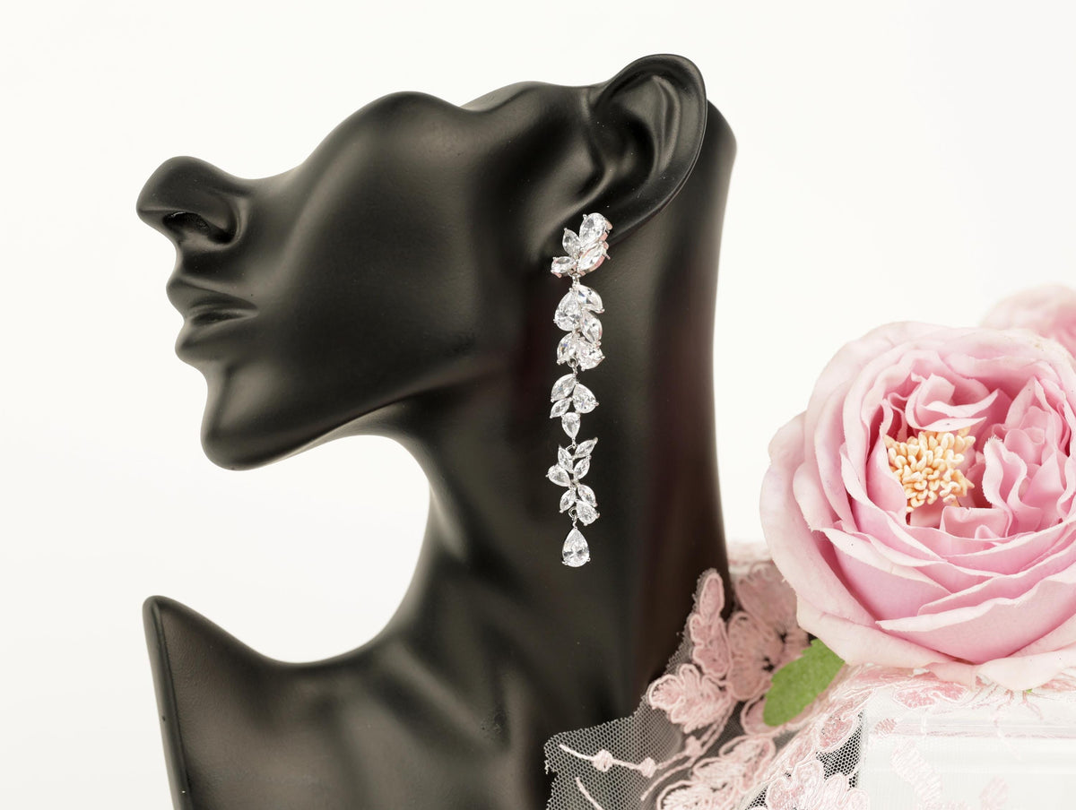 A black mannequin head wearing a pair of diamond earrings with a long, dangling design, accompanied by a pink rose.