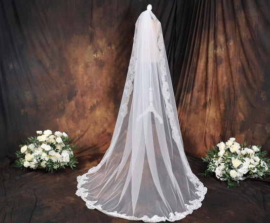 A white wedding veil with lace trim is displayed in front of a brown background, accompanied by two bouquets of white flowers.
