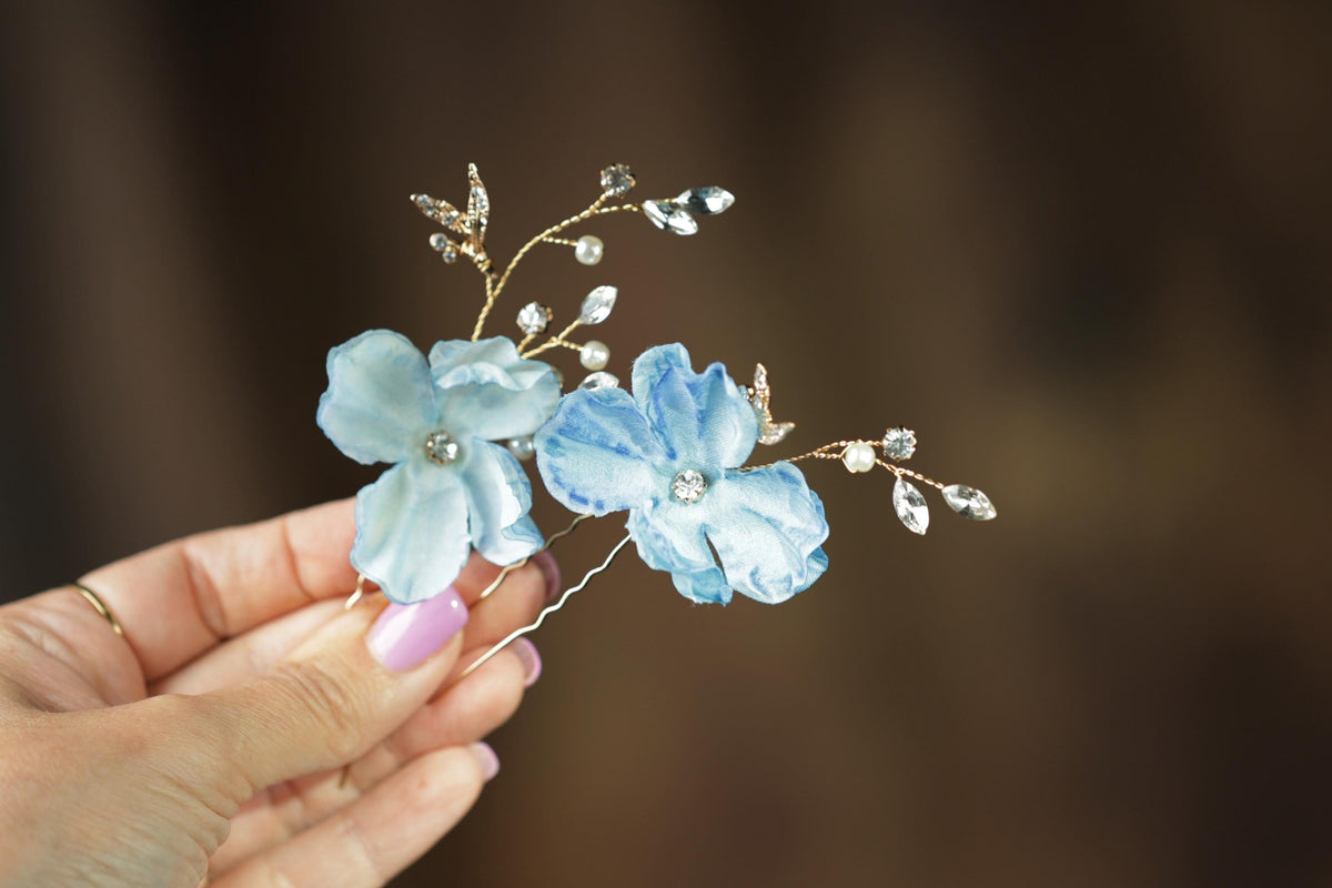 A hand holding a pair of blue flower-shaped hair clips with sparkling crystal embellishments.
