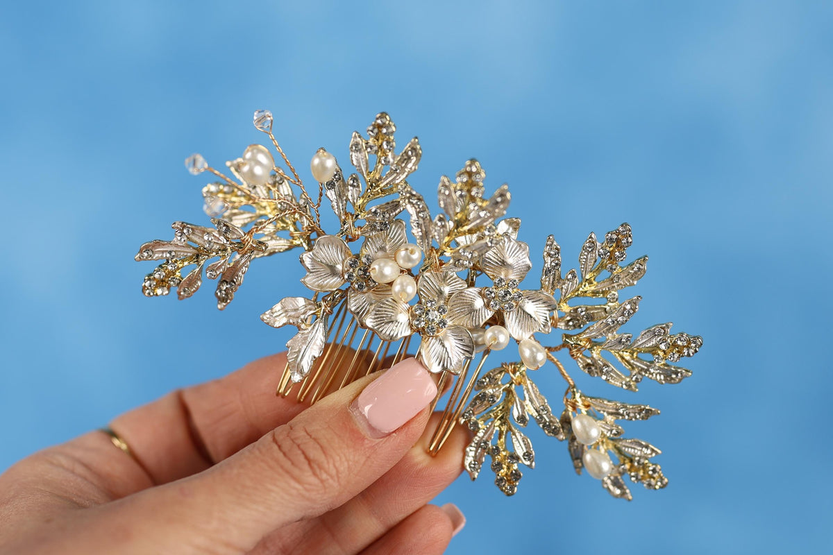 A hand holding a decorative hair accessory with gold and white floral elements against a blue background.