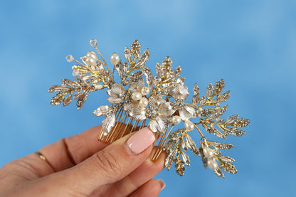 A hand holding a decorative hair accessory with gold and white floral elements against a blue background.
