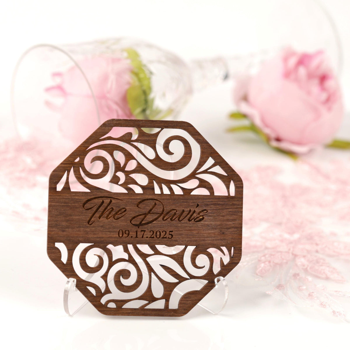 A wooden coaster with a floral design and the name "The Davis" engraved on it, placed on a white surface with a pink rose and a wine glass in the background.